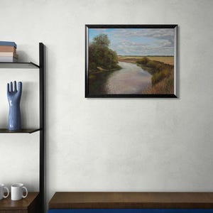 Puede incluir: Una pintura de paisaje enmarcada representa un r&iacute;o que serpentea a trav&eacute;s de un campo bajo un cielo nublado. La obra de arte tiene un marco negro y se exhibe en una pared blanca. Una escultura de mano de cer&aacute;mica azul y una estanter&iacute;a con libros y tazas son visibles.