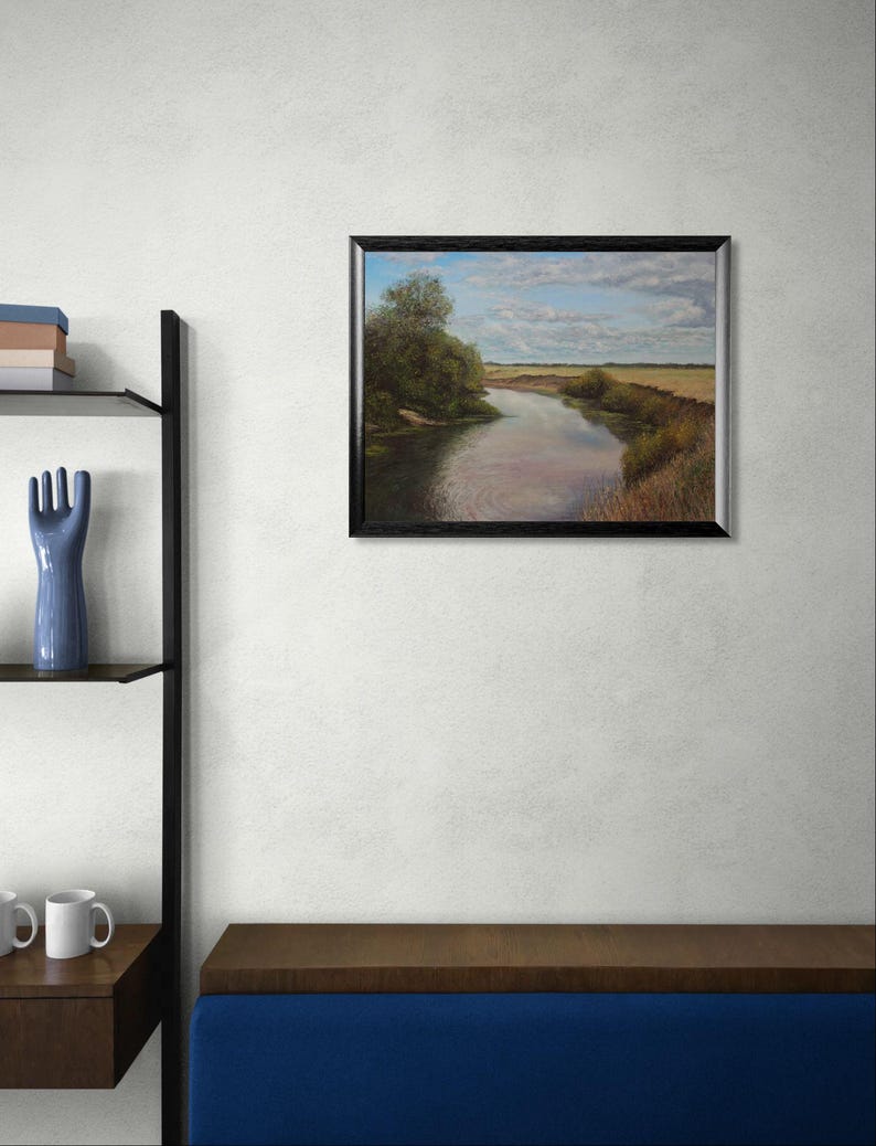 Puede incluir: Una pintura de paisaje enmarcada representa un r&iacute;o que serpentea a trav&eacute;s de un campo bajo un cielo nublado. La obra de arte tiene un marco negro y se exhibe en una pared blanca. Una escultura de mano de cer&aacute;mica azul y una estanter&iacute;a con libros y tazas son visibles.