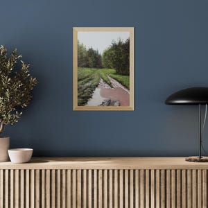 May include: A framed landscape print depicts a muddy path through a forest, with green trees and a reflective puddle. The artwork is in a light wood frame, displayed on a blue wall. A potted plant, a small bowl, and a black lamp are also visible.
