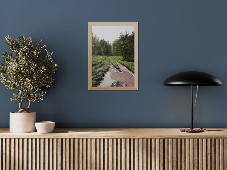 May include: A framed landscape print depicts a muddy path through a forest, with green trees and a reflective puddle. The artwork is in a light wood frame, displayed on a blue wall. A potted plant, a small bowl, and a black lamp are also visible.