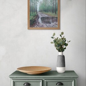 May include: A framed landscape painting of a road through a forest with a puddle. The painting is displayed above a green chest of drawers with black handles. A wooden bowl and a vase with greenery are on the chest.