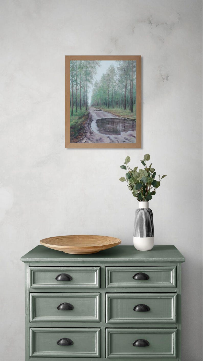 May include: A framed landscape painting of a road through a forest with a puddle. The painting is displayed above a green chest of drawers with black handles. A wooden bowl and a vase with greenery are on the chest.