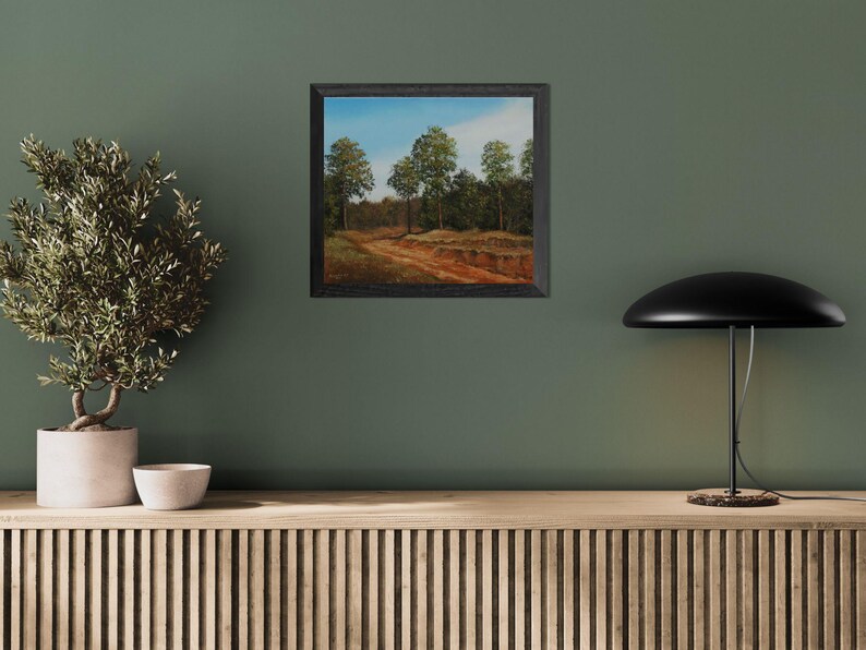 Pu&ograve; includere: Un dipinto di paesaggio incorniciato raffigura un sentiero sterrato che si snoda attraverso una foresta di alberi verdi sotto un cielo blu. Il dipinto &egrave; in una cornice nera ed &egrave; appeso a una parete verde. Una piccola pianta in vaso e una lampada sono in primo piano.