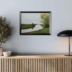 Pu&ograve; includere: Un dipinto di paesaggio incorniciato raffigura una serena scena fluviale sotto un cielo nuvoloso. L'opera d'arte, in una cornice nera, mostra un fiume che si snoda attraverso rive e alberi verdi. Il dipinto &egrave; appeso a una parete azzurra sopra un mobile in legno.