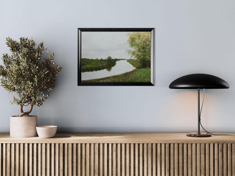 Pu&ograve; includere: Un dipinto di paesaggio incorniciato raffigura una serena scena fluviale sotto un cielo nuvoloso. L'opera d'arte, in una cornice nera, mostra un fiume che si snoda attraverso rive e alberi verdi. Il dipinto &egrave; appeso a una parete azzurra sopra un mobile in legno.