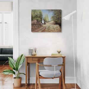 May include: An oil painting depicts a dirt path winding through a forest, with trees lining both sides under a blue sky. The artwork is displayed on a white wall above a wooden desk and a gray chair. A floor lamp and potted plant are also visible.