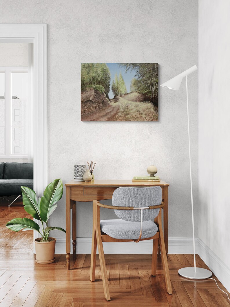 May include: An oil painting depicts a dirt path winding through a forest, with trees lining both sides under a blue sky. The artwork is displayed on a white wall above a wooden desk and a gray chair. A floor lamp and potted plant are also visible.