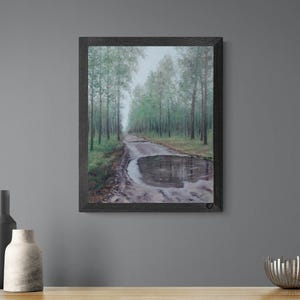 May include: Framed artwork depicting a muddy road with a large puddle reflecting the sky. The road is flanked by tall green trees. The artwork is presented in a dark wooden frame.