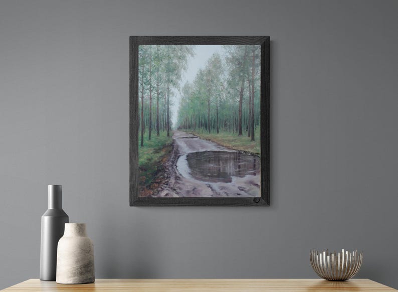 May include: Framed artwork depicting a muddy road with a large puddle reflecting the sky. The road is flanked by tall green trees. The artwork is presented in a dark wooden frame.