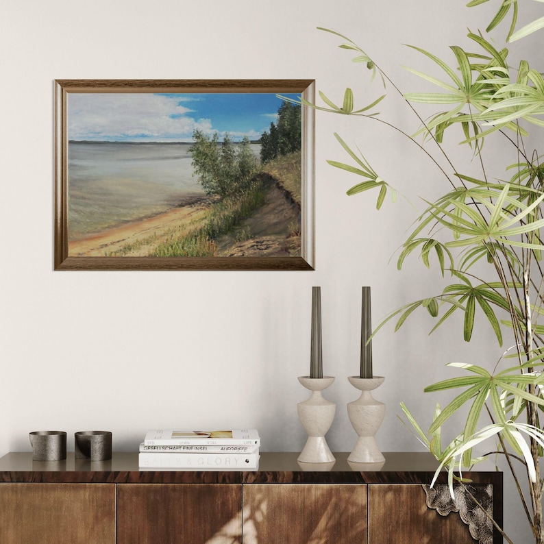 K&ouml;nnte beinhalten: Ein gerahmtes Landschaftsgem&auml;lde, das eine ruhige Szene am Wasser zeigt. Das Kunstwerk zeigt einen Sandstrand, &uuml;ppiges Gr&uuml;n und eine weite Wasserfl&auml;che unter einem blauen Himmel mit flauschigen Wolken. Der Rahmen hat eine warme braune Farbe.