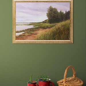 May include: A framed landscape painting shows a tranquil waterside scene with a sandy path, tall grasses, and trees under a cloudy sky. Below, a decorative tray holds figs, red mugs with plants, and a woven basket.