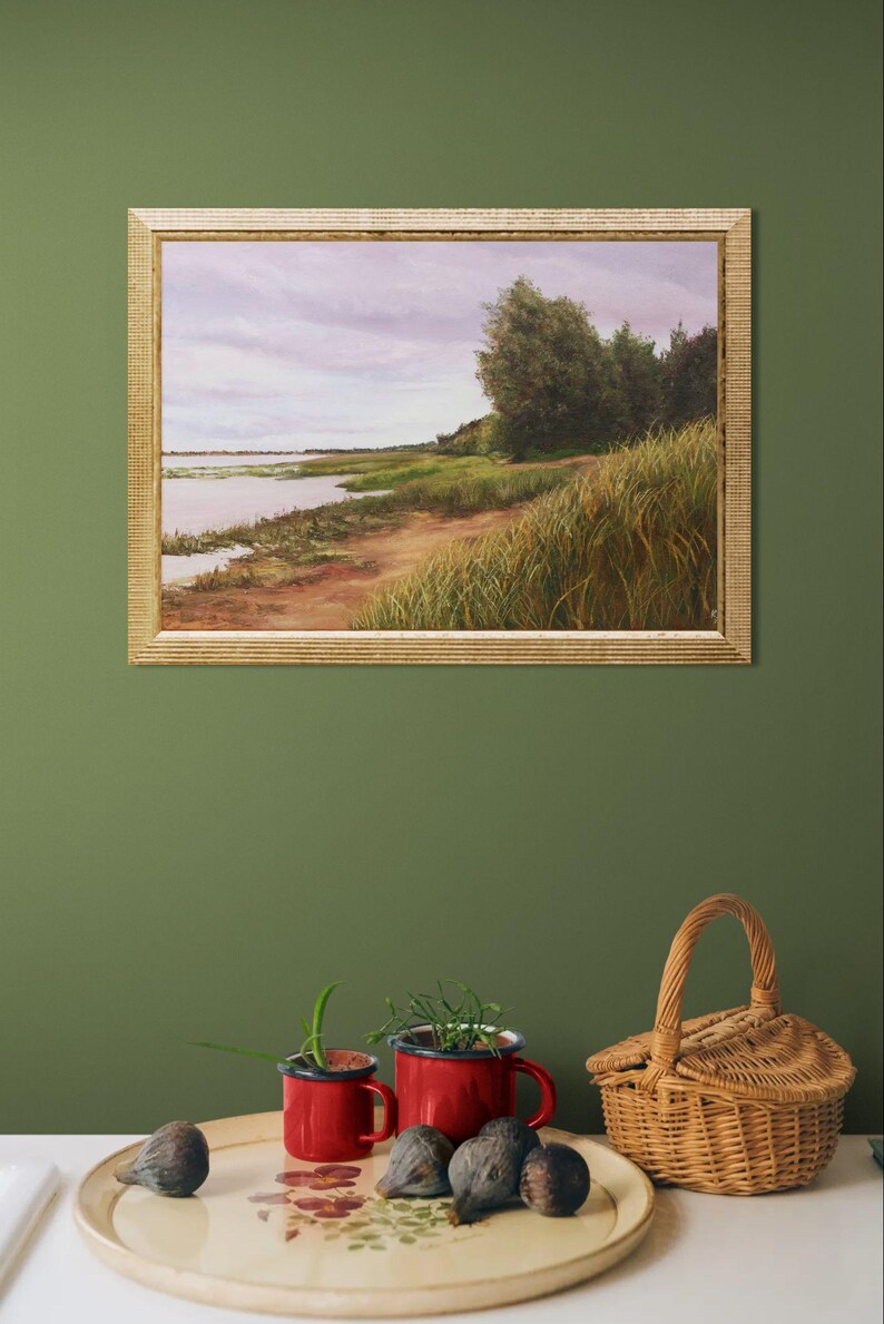 May include: A framed landscape painting shows a tranquil waterside scene with a sandy path, tall grasses, and trees under a cloudy sky. Below, a decorative tray holds figs, red mugs with plants, and a woven basket.