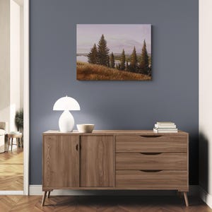 May include: A landscape painting depicting a serene scene with evergreen trees, a body of water, and a field of golden grass. The artwork is displayed on a wall above a wooden cabinet with a white lamp and a decorative bowl.