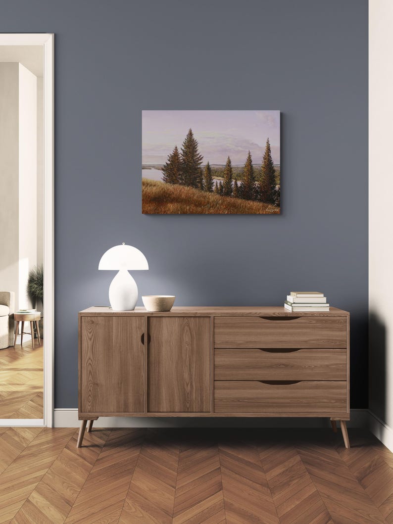 May include: A landscape painting depicting a serene scene with evergreen trees, a body of water, and a field of golden grass. The artwork is displayed on a wall above a wooden cabinet with a white lamp and a decorative bowl.