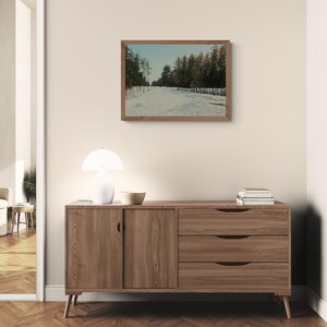 May include: A framed landscape painting depicts a snow-covered path winding through a forest of evergreen trees. The artwork hangs above a wooden sideboard with two drawers and a cabinet door. A white lamp and a wooden bowl sit on top.