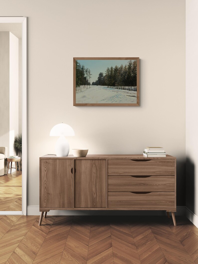 May include: A framed landscape painting depicts a snow-covered path winding through a forest of evergreen trees. The artwork hangs above a wooden sideboard with two drawers and a cabinet door. A white lamp and a wooden bowl sit on top.