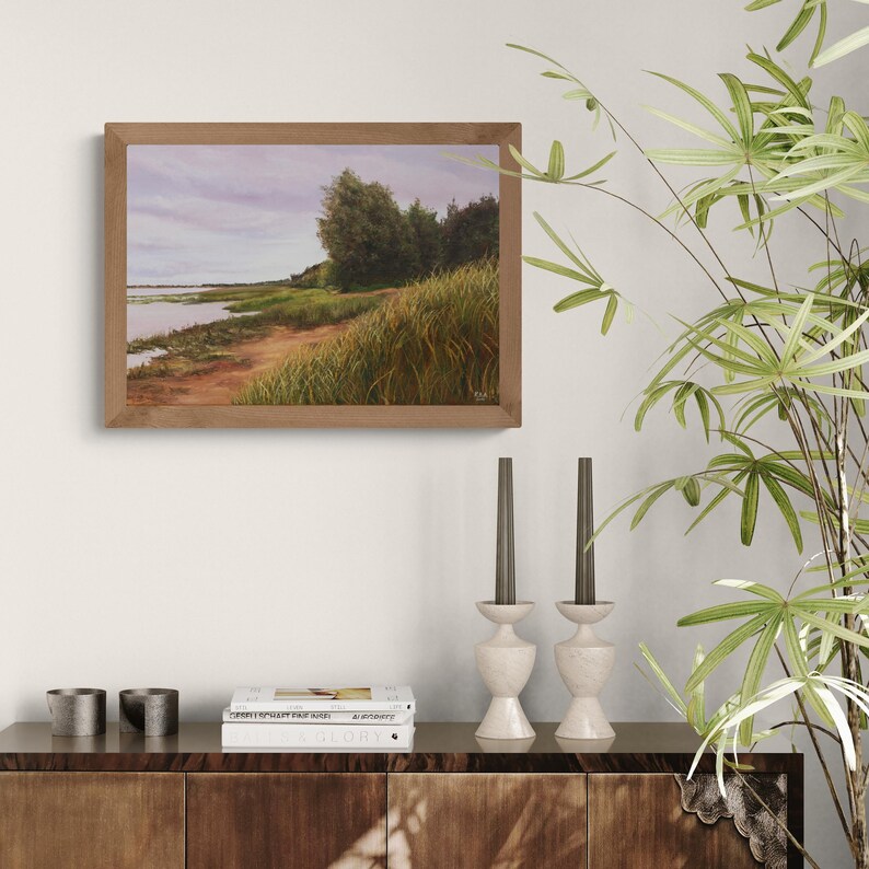 May include: A framed landscape painting showing a waterside scene. The artwork features a sandy bank, tall grasses, and trees under a cloudy sky. The frame is a warm, natural wood tone, complementing the earthy colours of the painting.