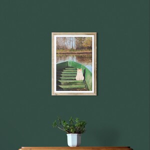 May include: A framed print of a green boat on a lake with a cat sitting in it. The boat is in the foreground, and the lake and trees are in the background. The print is hanging on a dark green wall above a wooden dresser.
