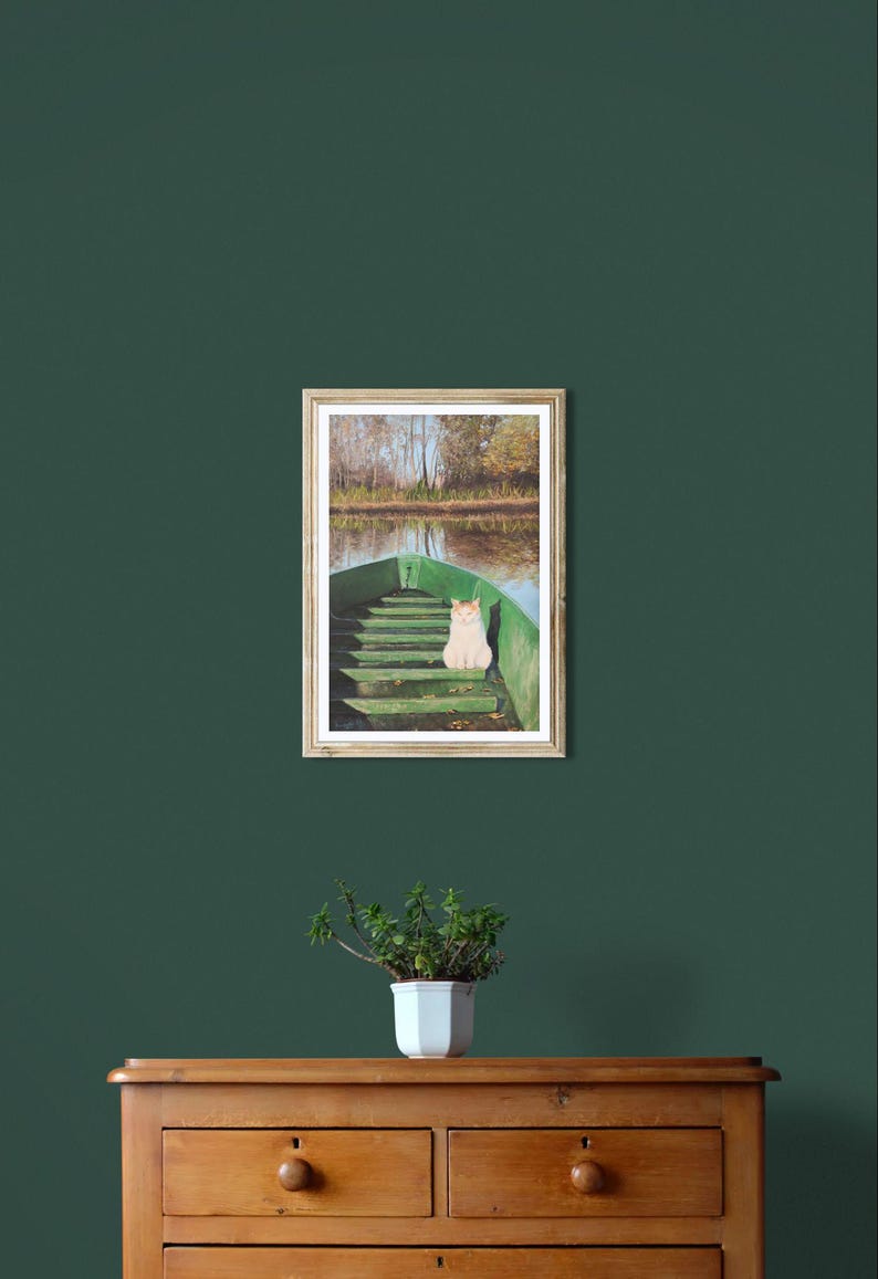May include: A framed print of a green boat on a lake with a cat sitting in it. The boat is in the foreground, and the lake and trees are in the background. The print is hanging on a dark green wall above a wooden dresser.