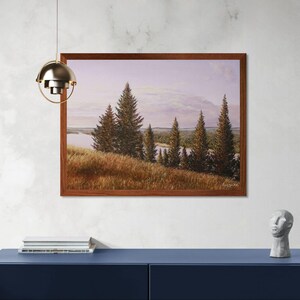 May include: A framed landscape painting depicts a serene scene with evergreen trees, a grassy hillside, and a body of water under a soft, cloudy sky. The artwork is encased in a dark brown wooden frame and hangs on a white wall above a blue cabinet.