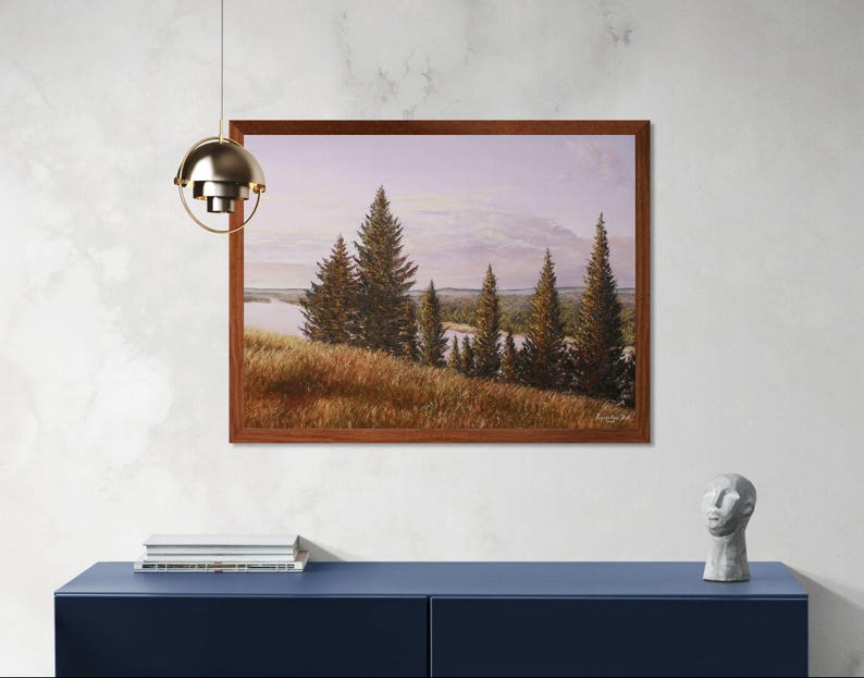 May include: A framed landscape painting depicts a serene scene with evergreen trees, a grassy hillside, and a body of water under a soft, cloudy sky. The artwork is encased in a dark brown wooden frame and hangs on a white wall above a blue cabinet.