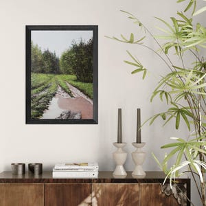 May include: A framed landscape print featuring a muddy dirt road winding through a forest. The road is flanked by green grass and trees, with a puddle in the foreground. The frame is black.
