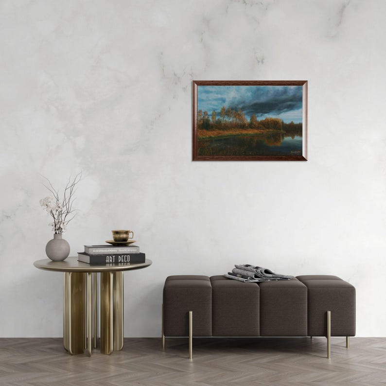 Pu&ograve; includere: Un dipinto di paesaggio incorniciato raffigura un sereno paesaggio acquatico sotto un cielo drammatico e nuvoloso. L'opera d'arte, con una cornice in legno scuro, presenta un lago riflettente, alberi e una tavolozza di colori cupi di blu, marroni e arancioni. Il dipinto &egrave; esposto su una parete bianca.