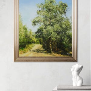 May include: Framed landscape painting depicting a tree-lined path under a blue sky. The artwork is in a gold-toned frame. A white sculpture of a torso and two books are on a wooden table.