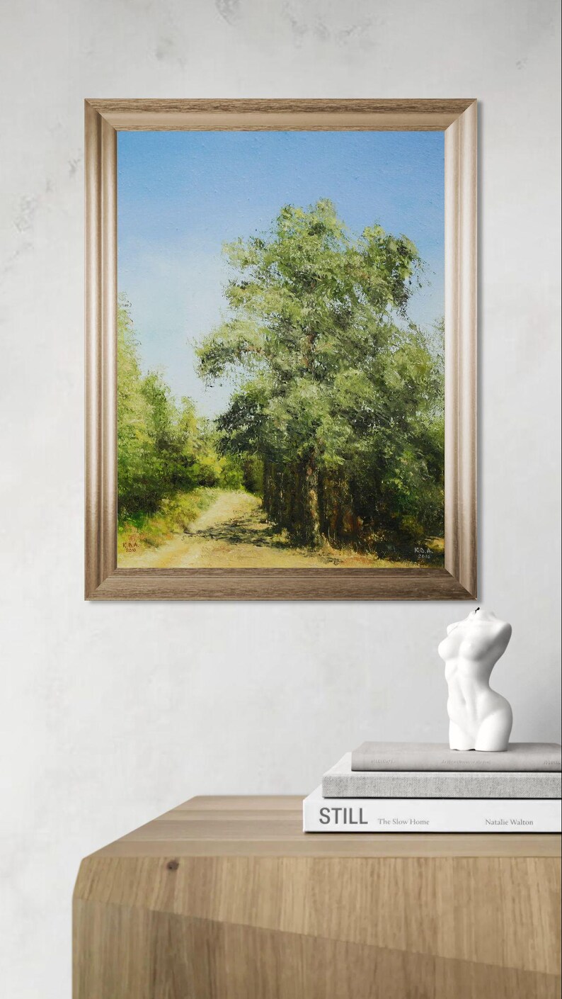 May include: Framed landscape painting depicting a tree-lined path under a blue sky. The artwork is in a gold-toned frame. A white sculpture of a torso and two books are on a wooden table.