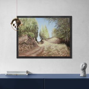 May include: A framed landscape painting depicts a dirt road winding through a forest. The artwork features a path with grassy areas and trees on either side, under a blue sky. The painting is displayed on a white wall above a blue cabinet.