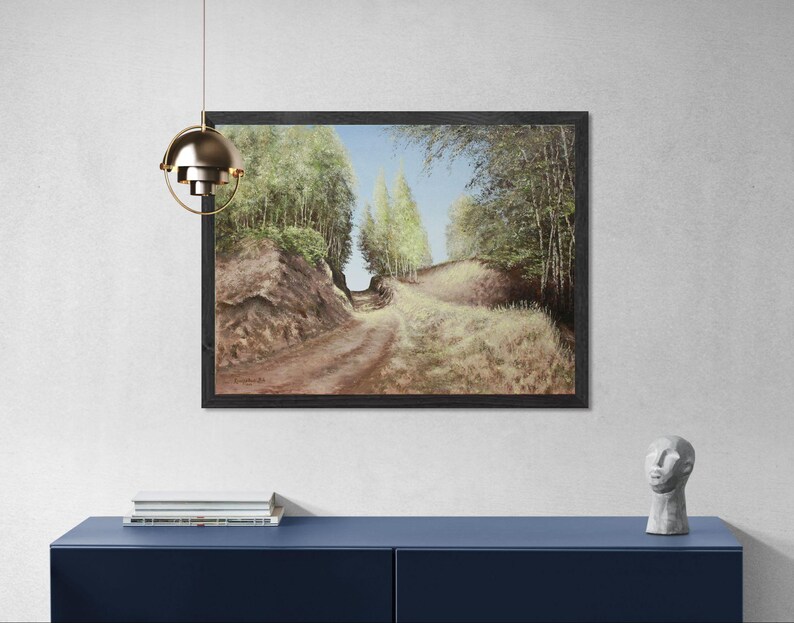 May include: A framed landscape painting depicts a dirt road winding through a forest. The artwork features a path with grassy areas and trees on either side, under a blue sky. The painting is displayed on a white wall above a blue cabinet.