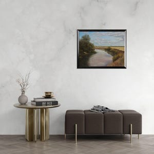 Puede incluir: Una pintura al &oacute;leo enmarcada de un paisaje fluvial cuelga de una pared blanca. La obra representa un r&iacute;o que serpentea a trav&eacute;s de campos y &aacute;rboles bajo un cielo nublado. Una peque&ntilde;a mesa auxiliar redonda con un jarr&oacute;n y libros est&aacute; cerca.