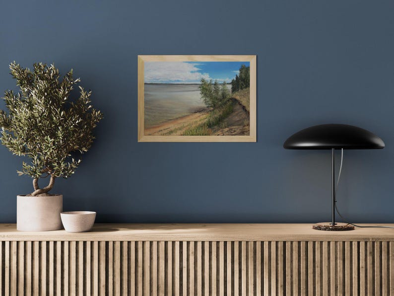 K&ouml;nnte beinhalten: Gerahmtes Landschaftsgem&auml;lde, das eine Szene am Wasser zeigt. Das Kunstwerk zeigt einen sandigen Uferbereich, &uuml;ppige gr&uuml;ne Vegetation und ein Gew&auml;sser unter einem bew&ouml;lkten Himmel mit blauen Flecken. Das Gem&auml;lde befindet sich in einem Holzrahmen.