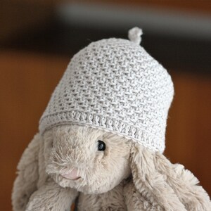 Könnte beinhalten: Eine beige Häkelmütze mit einem strukturierten Stichmuster. Die Mütze sitzt auf einem plüschigen, hellbraunen Stofftier.