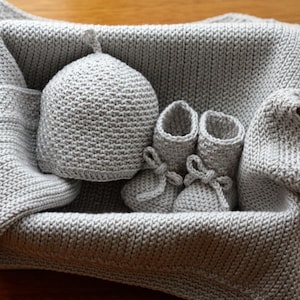 May include: A grey knitted baby blanket, hat and booties set. The items are arranged in a basket.