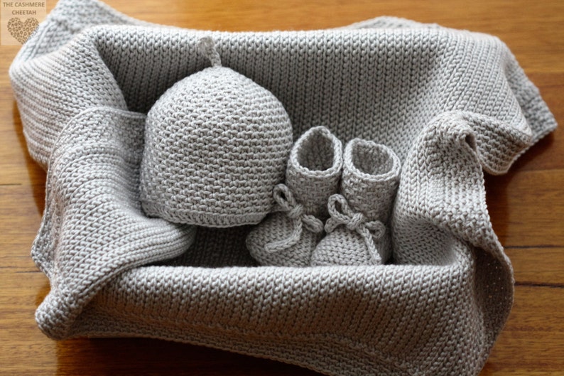 May include: A grey knitted baby blanket, hat and booties set. The items are arranged in a basket.