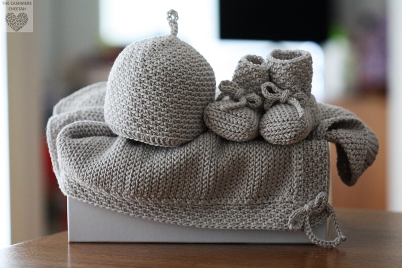 May include: A set of knitted baby clothes in a light grey colour. The set includes a hat, booties, and a cardigan. The items are arranged on a white box.