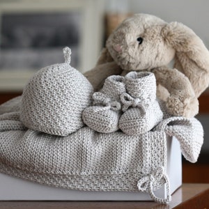 May include: A grey knitted baby blanket, hat and booties set in a white gift box with a plush bunny toy.