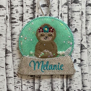 May include: A green and silver glitter snow globe ornament with a brown sloth wearing a flower crown inside. The base of the ornament has the name "Melanie" written in silver glitter.