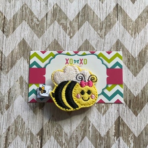 May include: A yellow and black embroidered bee with a pink bow and white wings. The bee is attached to a hair clip.