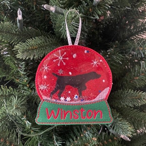 May include: A red and green Christmas ornament shaped like a snow globe with a black dog silhouette inside. The ornament has a white ribbon for hanging and the name "Winston" is embroidered on the base.