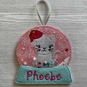 May include: Pink glitter snow globe ornament with a gray cat wearing a red Santa hat. The base of the ornament is light blue with the name "Phoebe" embroidered in pink.