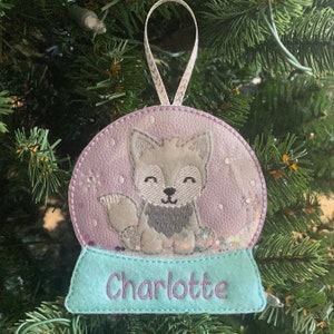 May include: A purple and white snow globe ornament with a gray fox inside. The base of the ornament is light blue with the name "Charlotte" embroidered on it.