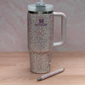 May include: A white stainless steel travel mug with a white lid and handle. The mug is covered in iridescent rhinestones. A matching rhinestone pen is lying next to the mug.