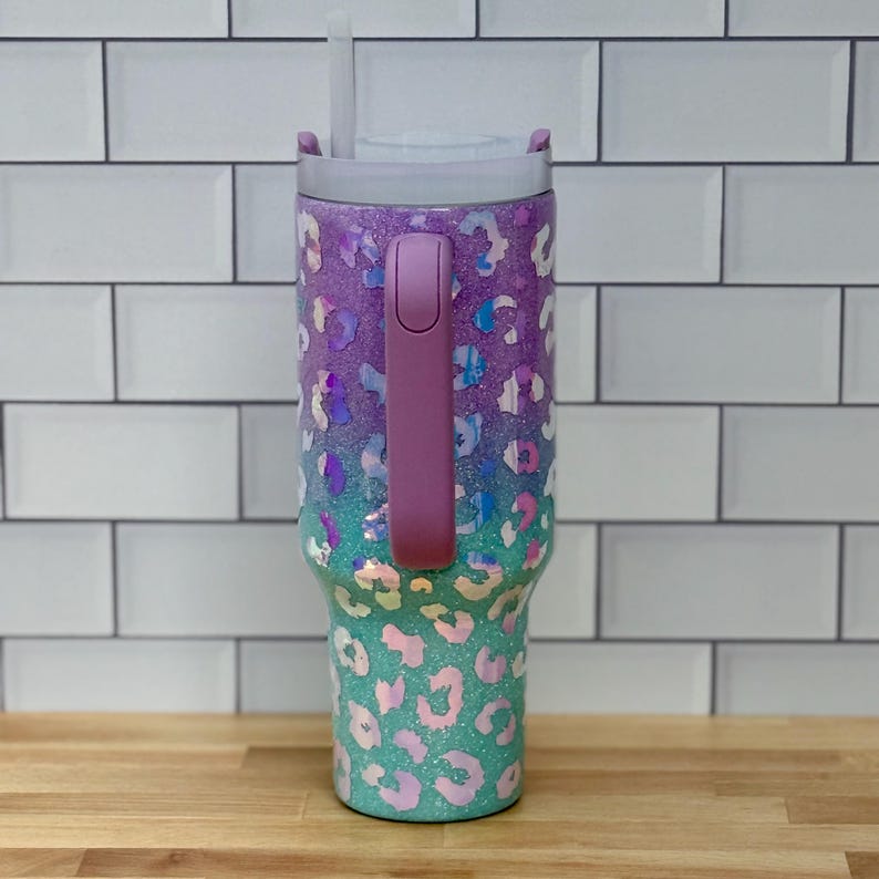 Puede incluir: Un vaso con purpurina, asa y tapa moradas. El vaso presenta un dise&ntilde;o de estampado de leopardo en tonos morados, turquesas e iridiscentes. Una pajita blanca est&aacute; insertada en la tapa. El vaso est&aacute; dise&ntilde;ado para mantener las bebidas fr&iacute;as.