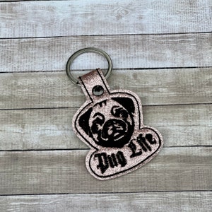 Peut inclure: Un porte-cl&eacute;s en paillettes roses avec une silhouette de carlin noire et le texte "Pug Life" en noir.