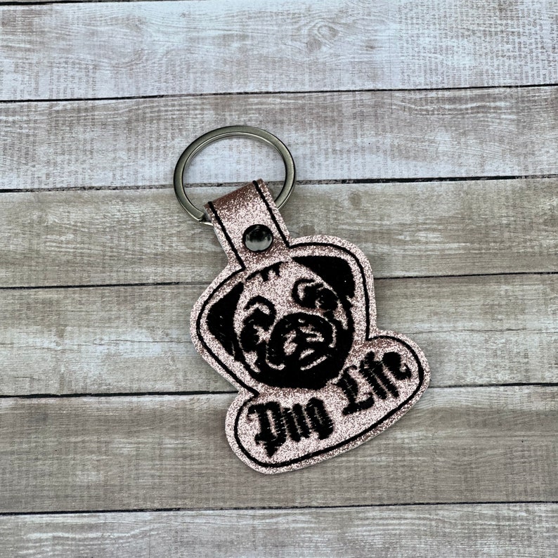 Peut inclure: Un porte-cl&eacute;s en paillettes roses avec une silhouette de carlin noire et le texte "Pug Life" en noir.