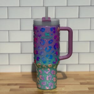 Puede incluir: Un vaso Stanley colorido con un dise&ntilde;o de estampado de leopardo. El vaso presenta un degradado de colores, incluyendo morado, azul y verde, con un asa y tapa moradas a juego. El logotipo de Stanley es visible en la parte delantera.