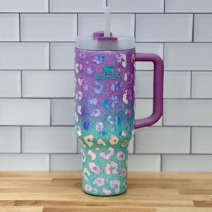 Puede incluir: Vaso Stanley con un dise&ntilde;o degradado en morado, azul y verde azulado. El vaso presenta un estampado de leopardo y un asa morada. Incluye una tapa transparente y una pajita blanca. El logotipo de Stanley es visible.