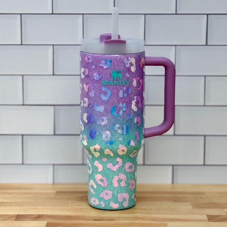 Puede incluir: Vaso Stanley con un dise&ntilde;o degradado en morado, azul y verde azulado. El vaso presenta un estampado de leopardo y un asa morada. Incluye una tapa transparente y una pajita blanca. El logotipo de Stanley es visible.